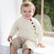 Baby wearing a cream-colored Aran sweater with three shoulder buttons and cable knit design, seated on a windowsill in light brown shorts – cozy and stylish knitwear for infants. Baby wearing a cream-colored Aran sweater with three shoulder buttons and cable knit design, seated on a windowsill in light brown shorts – cozy and stylish knitwear for infants.